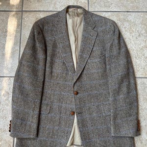 May include: A brown tweed blazer with a herringbone pattern and blue accents. The blazer has a notched lapel, two brown buttons, and a beige lining. The sleeves have three brown buttons. The blazer is laid flat on a tiled surface.
