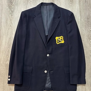 May include: A navy blue blazer featuring a notched lapel, two silver-toned buttons on the front, and two silver-toned buttons on each cuff. A yellow shield-shaped patch with the black text "SACHEM" is sewn onto the left chest pocket area.