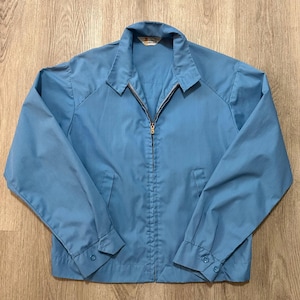 May include: A light blue zip-up jacket featuring a pointed collar, long sleeves with buttoned cuffs, and two front pockets. The garment has a central metal zipper closure and a small white brand label visible at the inner neckline area.