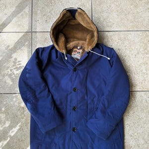 Puede incluir: Un abrigo largo azul marino con capucha forrada de piel sintética marrón. El abrigo tiene una botonadura frontal y un cordón en la capucha. El abrigo es de estilo vintage, adecuado para el clima frío.