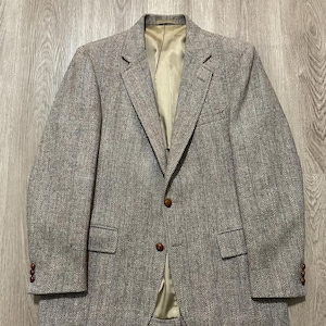 May include: A light brown tweed blazer with a herringbone pattern. The jacket features a notched lapel, two front pockets, and brown buttons. The interior lining is a light tan colour. A classic jacket suitable for various occasions.