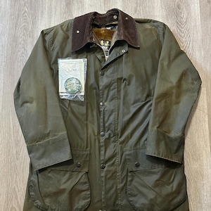 Puede incluir: Chaqueta de algodón encerado verde oliva con cuello de pana marrón. La chaqueta tiene cierre de cremallera y broches, múltiples bolsillos y una etiqueta de marca. Una prenda de abrigo clásica.