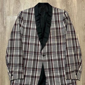 May include: A plaid blazer in a classic style. The jacket features a black satin lapel and a single-button closure. The plaid pattern includes burgundy, white, and black lines. The blazer is laid flat on a wooden surface.