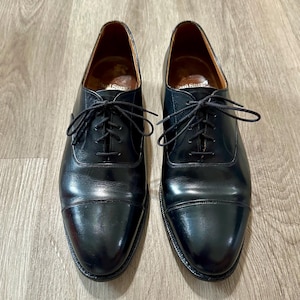 May include: A pair of black leather dress shoes with a cap toe design. The shoes have a lace-up closure and a polished finish. The interior of the shoes is a light brown color. The shoes are displayed on a light brown wooden surface.