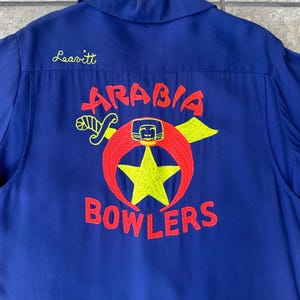May include: A blue bowling shirt with the name "Leavitt" embroidered in yellow script. The back features a red and yellow embroidered design with the words "Arabia Bowlers".
