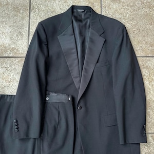 Op de afbeelding: Een formele zwarte smoking, bestaande uit een jasje met satijnen revers en een bijpassende broek. Het jasje heeft een sluiting met één knoop en manchetten met knopen. Het pak wordt gepresenteerd op een neutrale ondergrond.