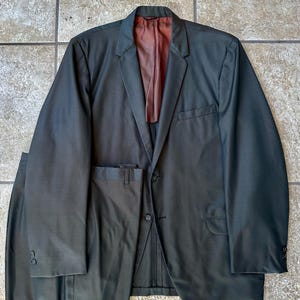 May include: A dark olive green suit set consisting of a single-breasted blazer with two buttons, notched lapels, and a chest pocket, paired with matching trousers. The blazer features a smooth, solid-colored dark reddish-brown interior lining.