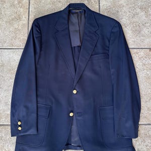 Puede incluir: Un blazer azul marino con botones dorados. El blazer tiene un diseño clásico con solapa de muesca, dos bolsillos delanteros y un bolsillo en el pecho. Las mangas tienen botones en los puños. El blazer está hecho de tela lisa.