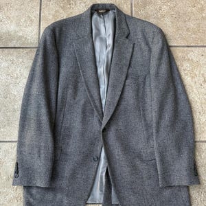 Puede incluir: Un blazer gris con estampado de espiga con cierre de dos botones y solapas con muescas. La chaqueta tiene un forro gris claro y dos bolsillos delanteros. Las mangas tienen tres botones en los puños. El blazer es de tejido texturizado.