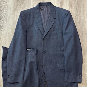 Puede incluir: Un traje gris carbón oscuro compuesto por una americana de botonadura sencilla y pantalones a juego. La americana presenta solapas de muesca, dos botones frontales, un bolsillo en el pecho, dos bolsillos con solapa y tres botones en cada puño.