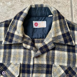 Puede incluir: Primer plano de una camisa de cuadros vintage en tonos grises, azul marino y amarillo. La camisa presenta un cuello, dos bolsillos en el pecho y cierres de botones. Una etiqueta con texto es visible cerca del cuello.