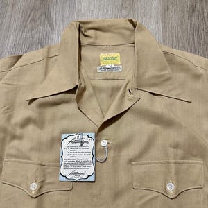 May include: A tan button-down shirt with a pointed collar, two chest pockets with buttoned flaps, and a size M tag. A light blue rectangular tag attached to the front reads "Austinized" with text detailing fabric care and shrinkage control.