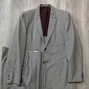 Puede incluir: Un traje de dos piezas que incluye una americana y pantalones a juego con un patrón de microcuadros en gris y negro. La americana tiene solapas de muesca, dos botones frontales y un forro interior de color burdeos oscuro. Puños con dos botones.