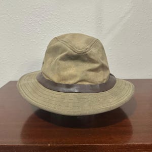 May include: A tan bucket hat featuring a structured crown and a wide brim with horizontal stitching. A dark brown band encircles the base of the crown, providing a contrast to the light tan fabric of the hat. The hat has a soft, worn texture.