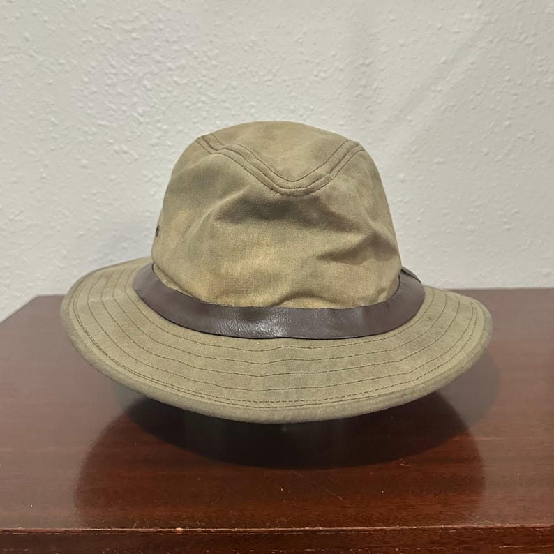 May include: A tan bucket hat featuring a structured crown and a wide brim with horizontal stitching. A dark brown band encircles the base of the crown, providing a contrast to the light tan fabric of the hat. The hat has a soft, worn texture.