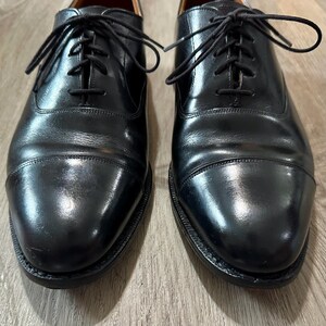 May include: A pair of black leather Oxford shoes with a cap toe. The shoes have black laces and a polished finish. The shoes are displayed on a wooden surface. The shoes are a classic style.