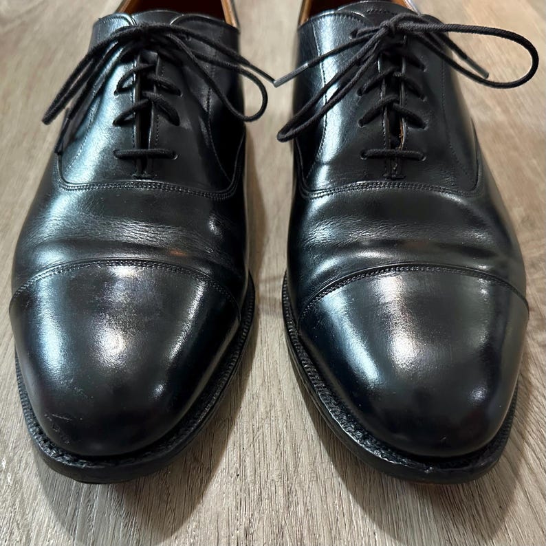 May include: A pair of black leather Oxford shoes with a cap toe. The shoes have black laces and a polished finish. The shoes are displayed on a wooden surface. The shoes are a classic style.