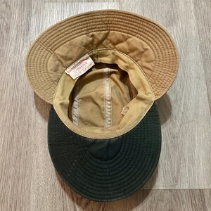 May include: A tan hat with a wide brim and a dark green underside. The interior features a white fabric tag with the text A Genuine Filson Garment C.C. Filson Co. Seattle, Wash. The hat has a structured crown and a stitched brim design.