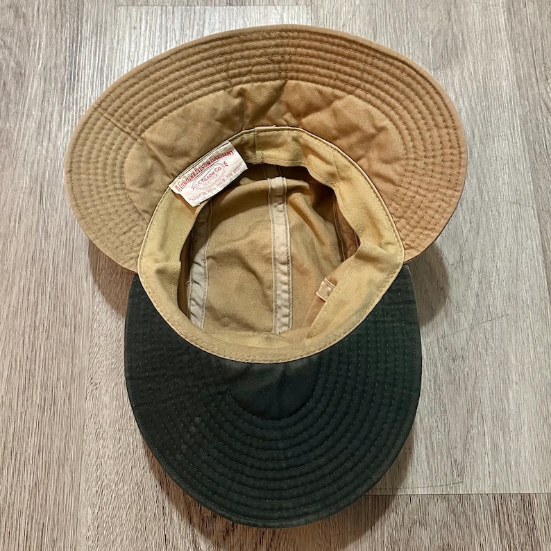 May include: A tan hat with a wide brim and a dark green underside. The interior features a white fabric tag with the text A Genuine Filson Garment C.C. Filson Co. Seattle, Wash. The hat has a structured crown and a stitched brim design.