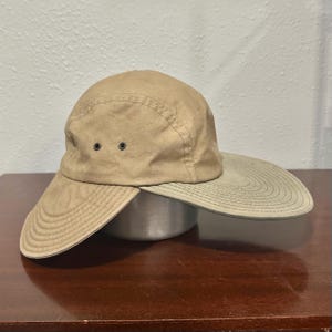 May include: A tan five-panel cap featuring a wide, extended brim that wraps around the sides. The hat is constructed from a solid-colored fabric and includes two small, dark metal ventilation eyelets on the side panel for airflow.