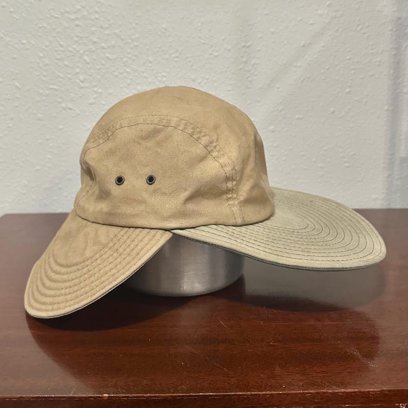 May include: A tan five-panel cap featuring a wide, extended brim that wraps around the sides. The hat is constructed from a solid-colored fabric and includes two small, dark metal ventilation eyelets on the side panel for airflow.