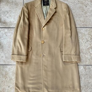 May include: A tan-colored, long coat with a classic design. The coat features a notched lapel, three buttons, and two front pockets. The coat has a label that reads "CAMERON PAUL".