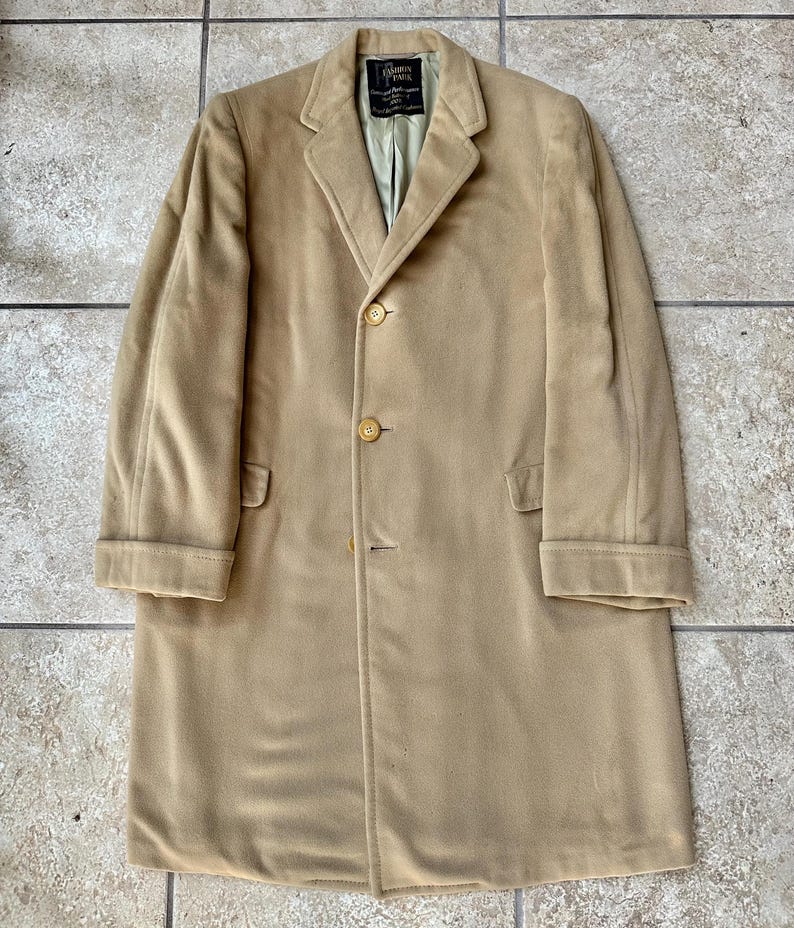 May include: A tan-colored, long coat with a classic design. The coat features a notched lapel, three buttons, and two front pockets. The coat has a label that reads "CAMERON PAUL".