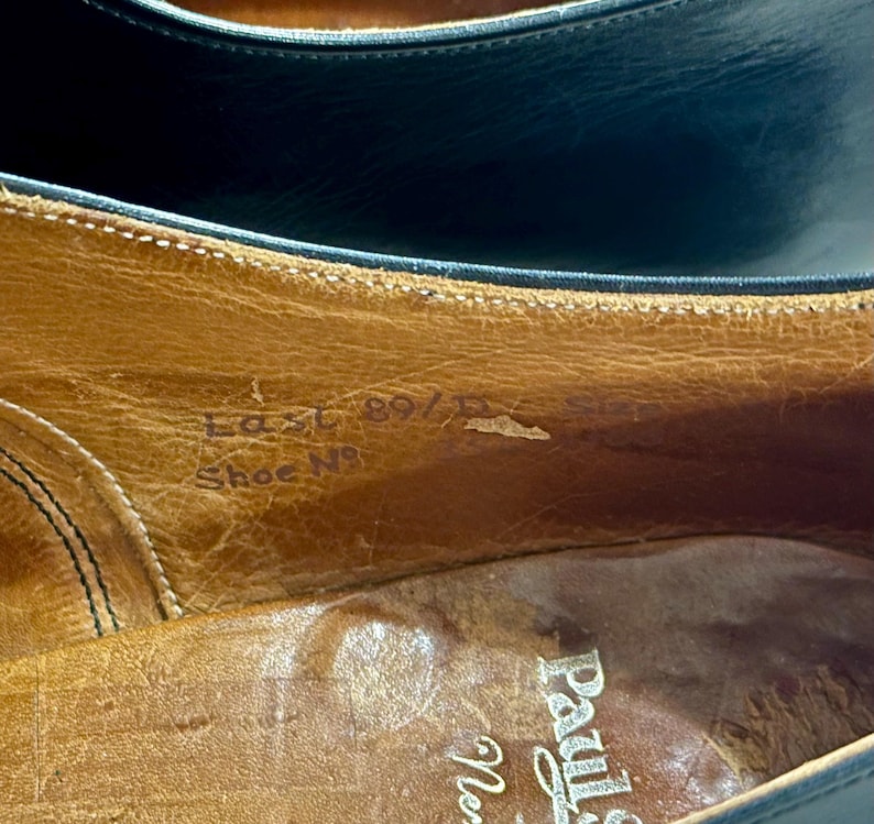 May include: Close-up of the interior of a leather shoe. The shoe's insole is a light brown colour, with visible text that reads "Last 89/D" and "Shoe No". The shoe's upper is a dark colour.