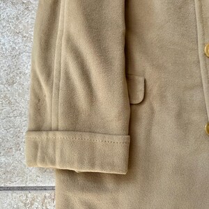 May include: Close-up of a tan coat, showcasing the sleeve with a folded cuff and a small pocket. The coat features a button closure and a classic design, suitable for outerwear.