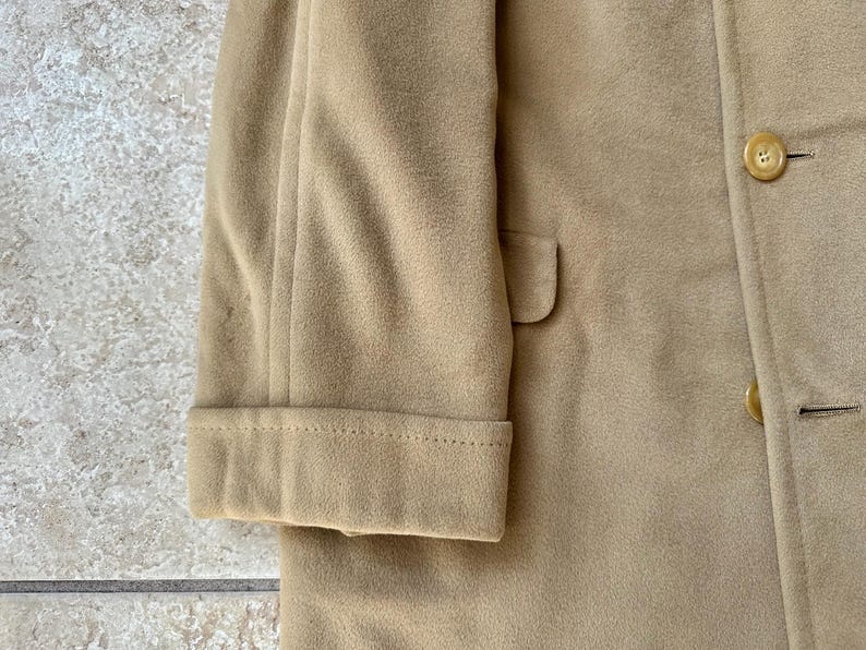 May include: Close-up of a tan coat, showcasing the sleeve with a folded cuff and a small pocket. The coat features a button closure and a classic design, suitable for outerwear.