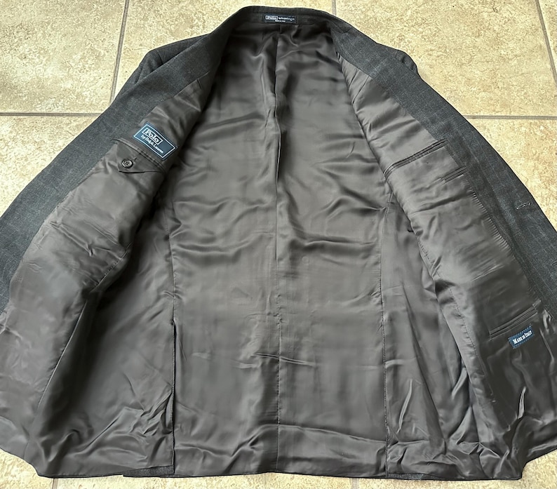 May include: A dark gray blazer with a pinstripe pattern. The blazer is unbuttoned and the lining is visible. The lining is a dark gray color and has a label that says "Polo Ralph Lauren".