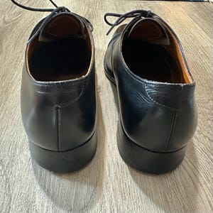 May include: A pair of black leather dress shoes with laces. The shoes have a classic design with a rounded toe and a low heel. The interior of the shoes is a light brown colour.