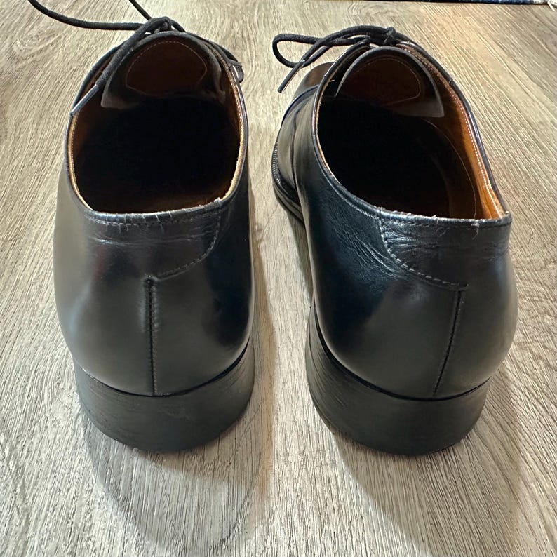 May include: A pair of black leather dress shoes with laces. The shoes have a classic design with a rounded toe and a low heel. The interior of the shoes is a light brown colour.