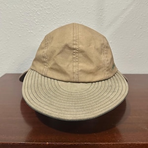 May include: A tan baseball cap featuring a five-panel construction with a curved, olive-toned brim. The brim displays concentric circular stitching patterns, and the crown is made of a smooth, solid-colored fabric with visible vertical seams.