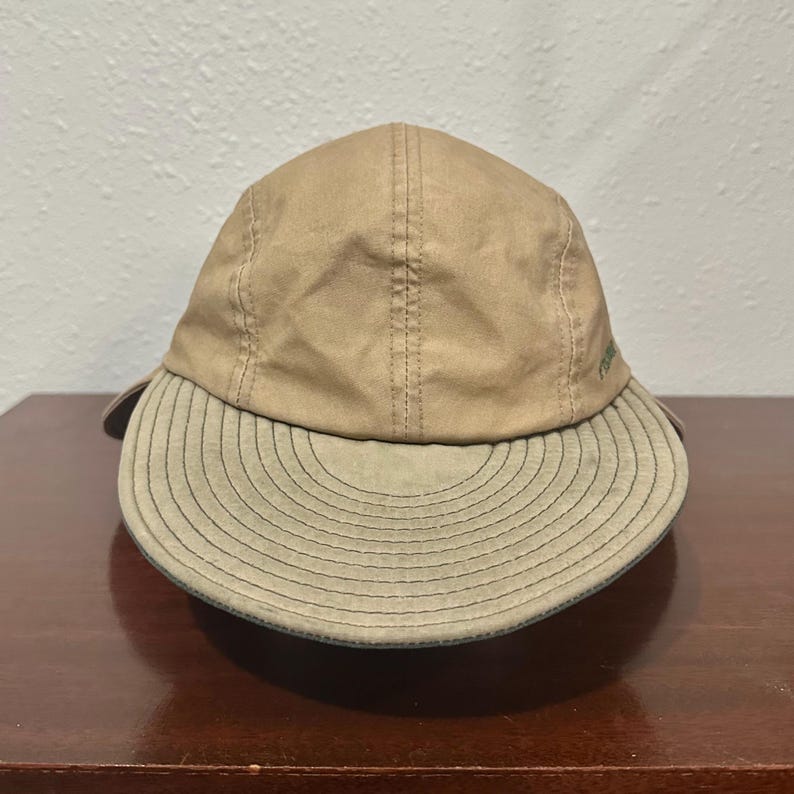 May include: A tan baseball cap featuring a five-panel construction with a curved, olive-toned brim. The brim displays concentric circular stitching patterns, and the crown is made of a smooth, solid-colored fabric with visible vertical seams.