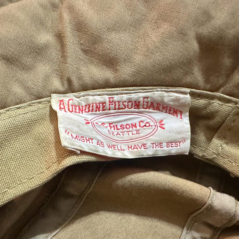 May include: A tan-colored hat featuring an interior white fabric label with red text that reads A GENUINE FILSON GARMENT, C.C. FILSON CO. SEATTLE, and MIGHT AS WELL HAVE THE BEST. The label is stitched onto the inner band of the tan fabric hat.