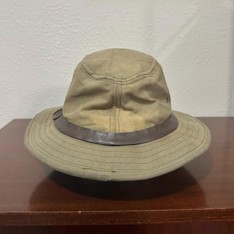 May include: A tan bucket hat featuring a dark brown leather band encircling the base of the crown. The hat is constructed with a rounded top, stitched seams, and a wide, downward-sloping brim that provides a structured shape and profile.