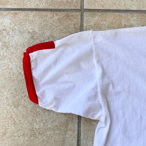 May include: A close-up view of a white t-shirt sleeve with a red cuff. The fabric appears soft and slightly creased. The sleeve is angled, revealing the shoulder seam.