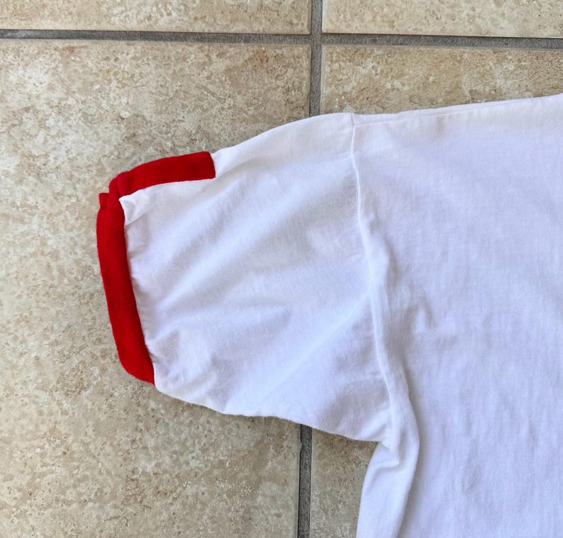 May include: A close-up view of a white t-shirt sleeve with a red cuff. The fabric appears soft and slightly creased. The sleeve is angled, revealing the shoulder seam.
