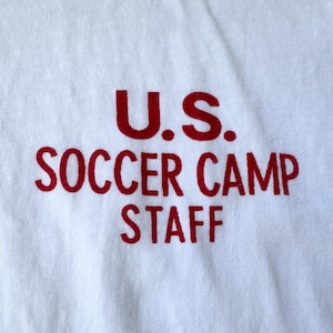 May include: A white t-shirt featuring the red text "U.S. SOCCER CAMP STAFF". The text is in a bold, sans-serif font. The shirt is made of a soft, cotton material. The text is printed across the front.