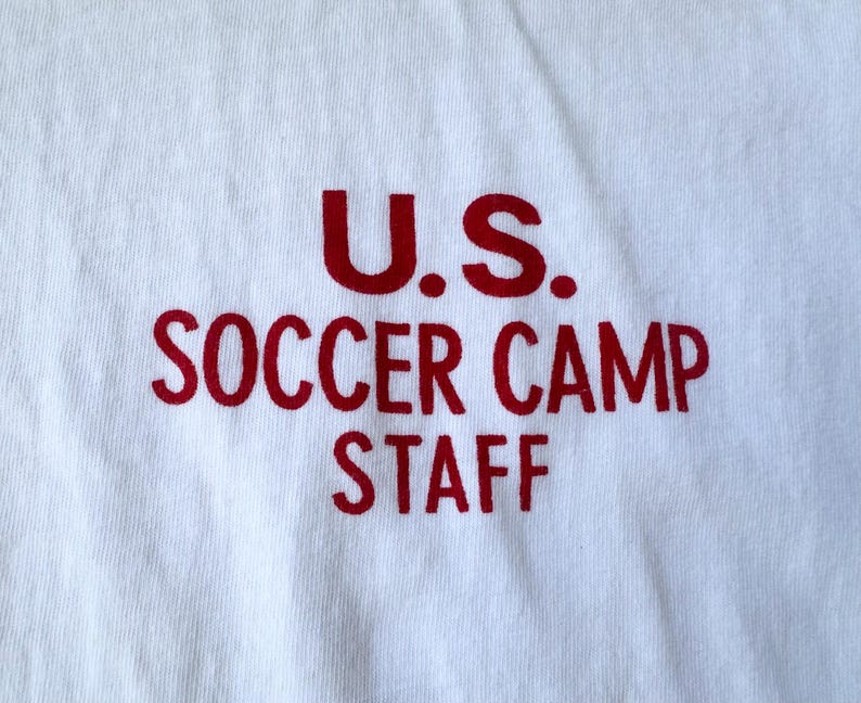 May include: A white t-shirt featuring the red text "U.S. SOCCER CAMP STAFF". The text is in a bold, sans-serif font. The shirt is made of a soft, cotton material. The text is printed across the front.