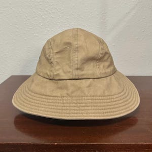 May include: A tan bucket hat with a rounded crown and a wide, downward-sloping brim. The hat features visible vertical stitching along the crown panels and multiple rows of circular stitching around the brim, providing a structured appearance.