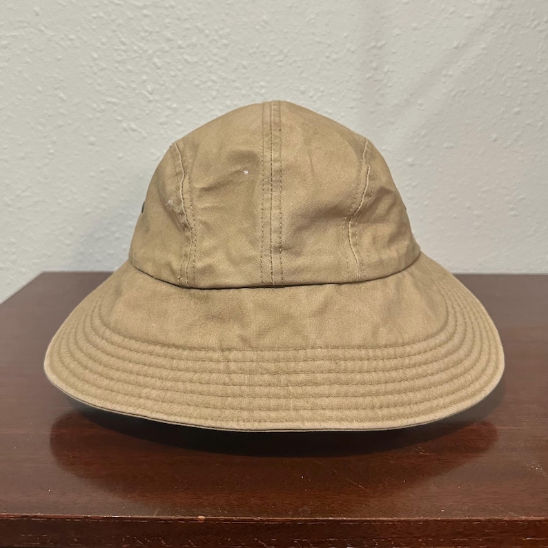 May include: A tan bucket hat with a rounded crown and a wide, downward-sloping brim. The hat features visible vertical stitching along the crown panels and multiple rows of circular stitching around the brim, providing a structured appearance.