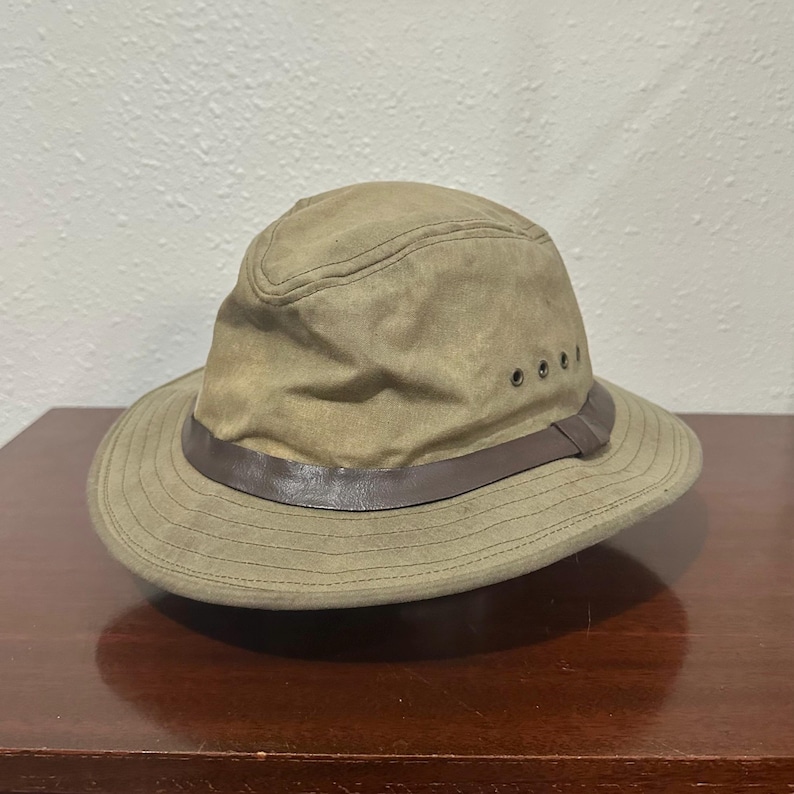 May include: A tan-coloured fedora-style hat featuring a dark brown leather band encircling the base of the crown. The hat is constructed from a matte fabric and includes three small metal ventilation eyelets on the side of the crown.
