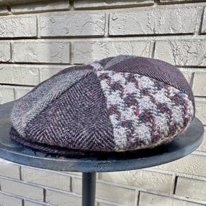May include: A patchwork newsboy cap in shades of gray, purple, and white. The cap features a herringbone pattern and a textured, woven design. The brim is a solid dark purple color.
