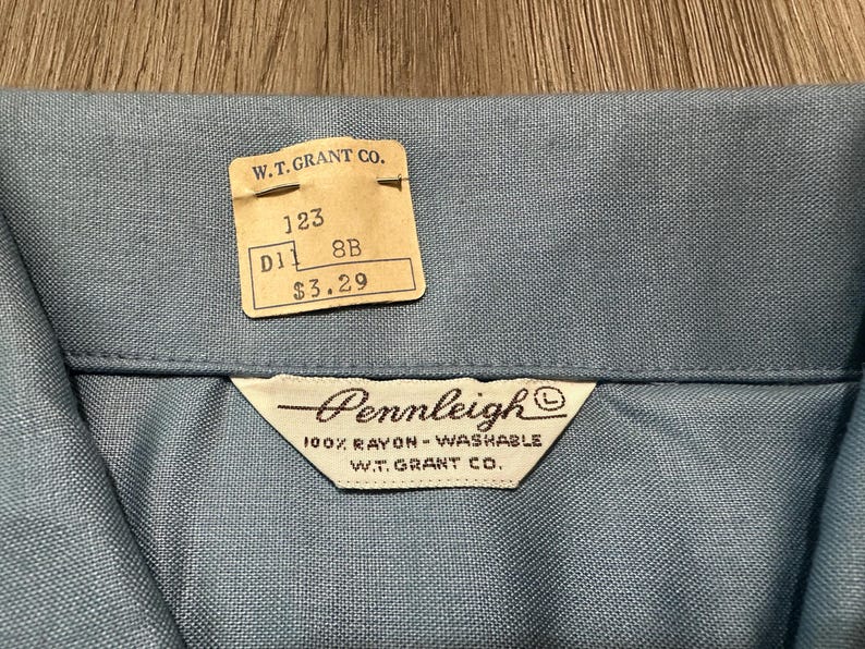 May include: A light blue shirt collar with a vintage W.T. Grant Co. paper price tag attached. The tag displays the text 123, D11, 8B, and $3.29. A white fabric label is sewn below the collar, reading Pennleigh, 100% Rayon-Washable, W.T. Grant Co.