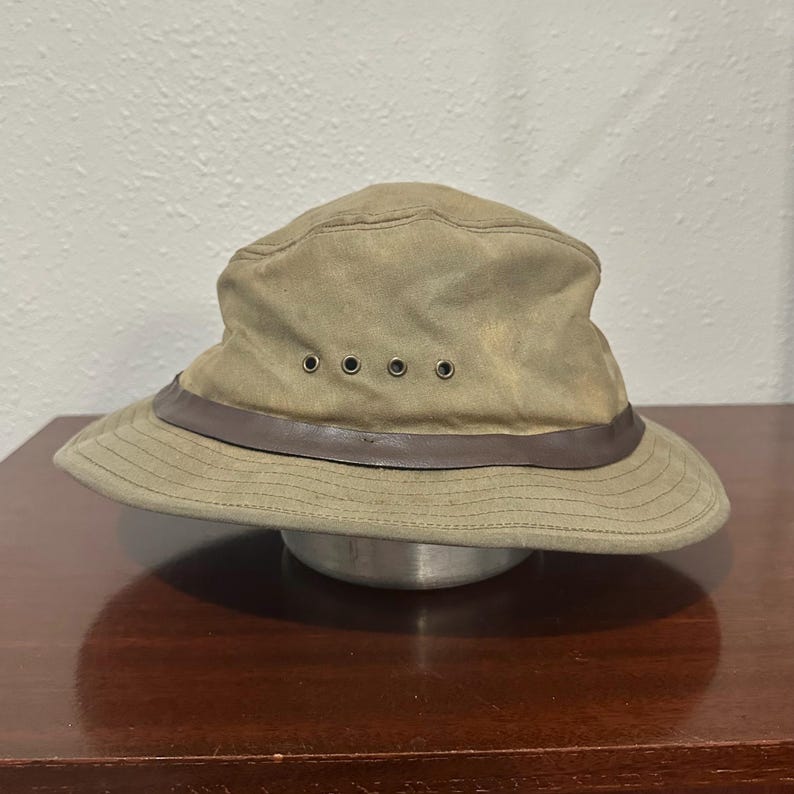 May include: A tan bucket hat featuring a dark brown leather band encircling the crown. The hat includes four small metal ventilation eyelets on the side and a wide, downward-sloping brim with visible stitching detail throughout the fabric.