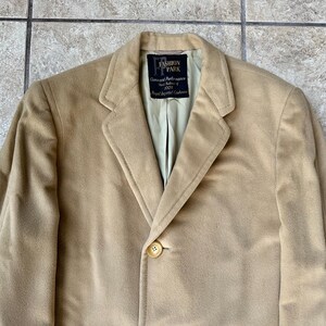 May include: A tan blazer with a single button closure. The blazer has a notched lapel and a light green lining. The label reads "FASHION PARK Guaranteed Performance 100% Royal Imported Cashmere."