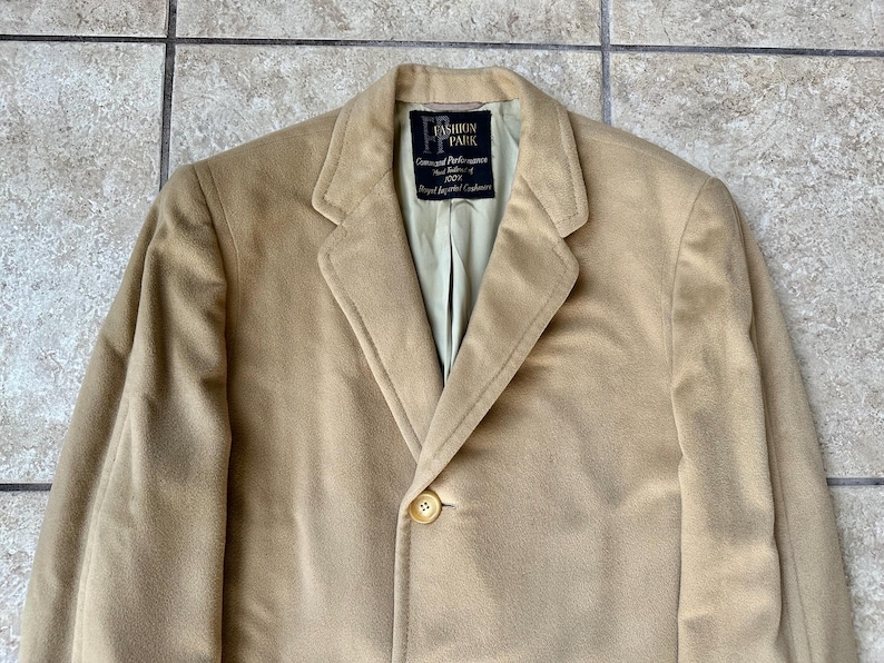 May include: A tan blazer with a single button closure. The blazer has a notched lapel and a light green lining. The label reads "FASHION PARK Guaranteed Performance 100% Royal Imported Cashmere."