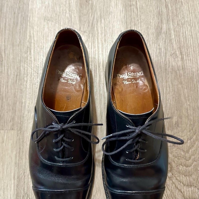 May include: A pair of black leather dress shoes with laces. The shoes have a classic design with a rounded toe and a polished finish. The interior of the shoes is a light brown colour, with the brand name "Paul Stuart New York" visible.